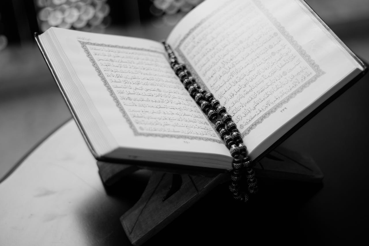 services-03 A black and white close-up of an open Quran on a wooden stand with prayer beads, symbolizing Islamic faith and devotion.